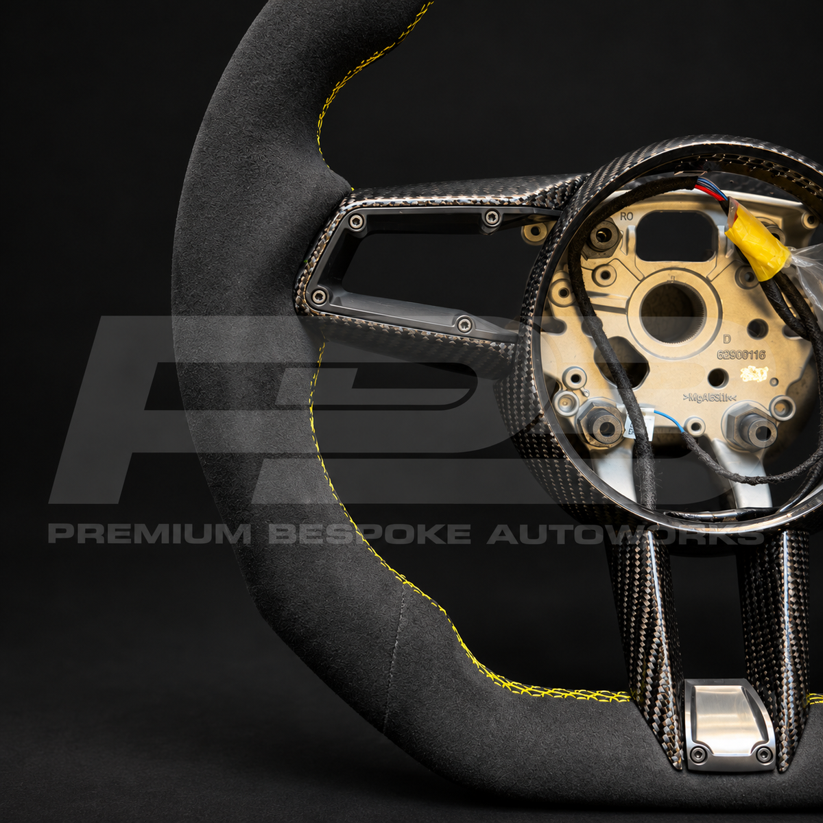 Porsche 911 992 custom steering wheel angled from the right, showing the black Alcantara grip, yellow stitching and carbon fibre trim