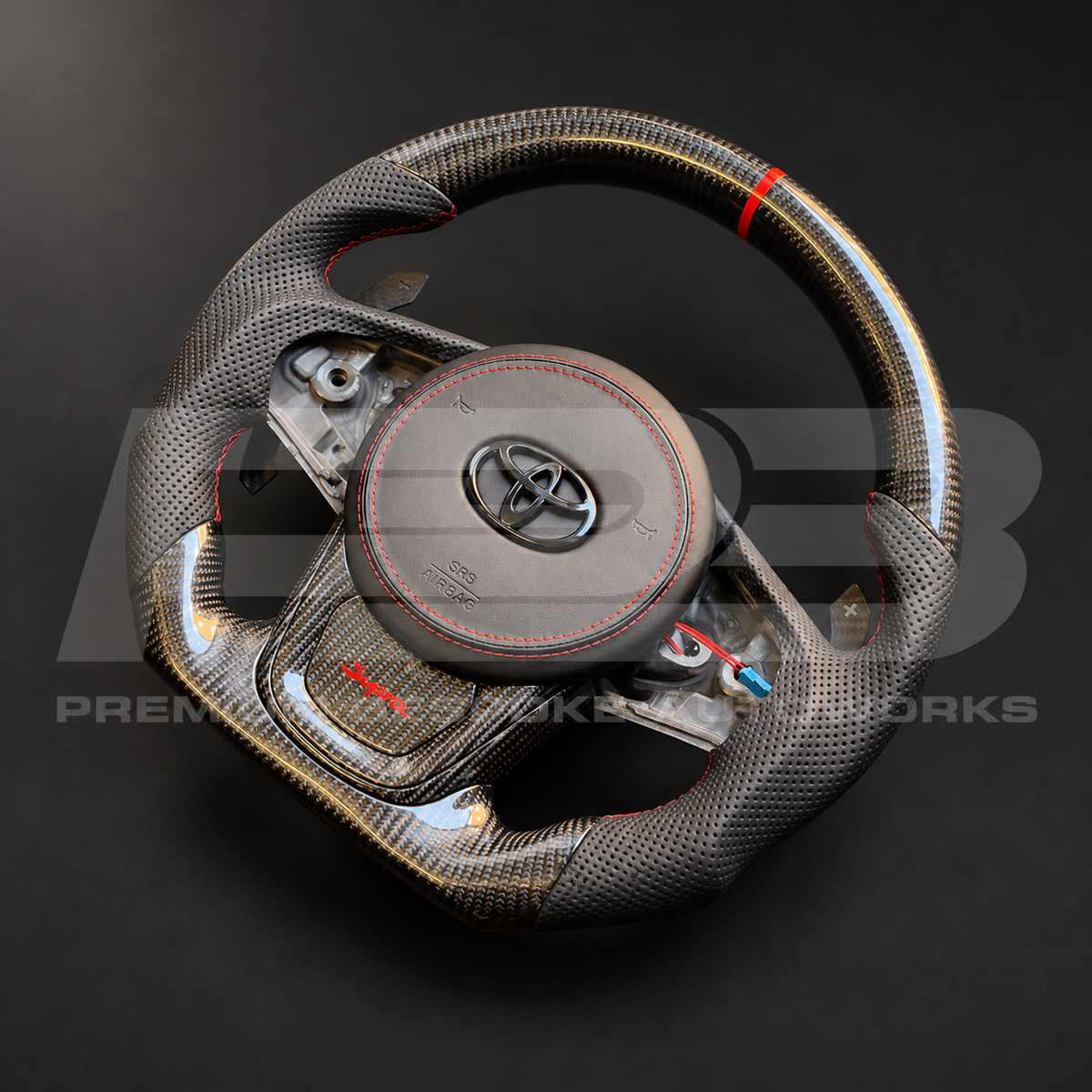 Toyota GR Supra MKV custom steering wheel from the opposite angle, gloss carbon fibre, perforated leather grips, red stitching and magnetic paddle shifters