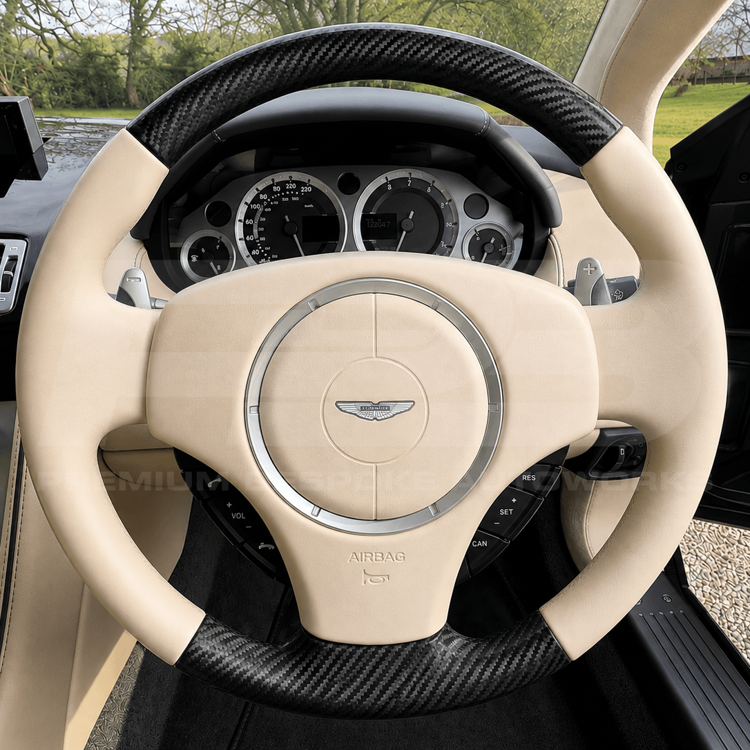 A close-up view of a luxury car's steering wheel featuring beige leather wrap, carbon fiber trim, and the Aston Martin wing logo in the center.