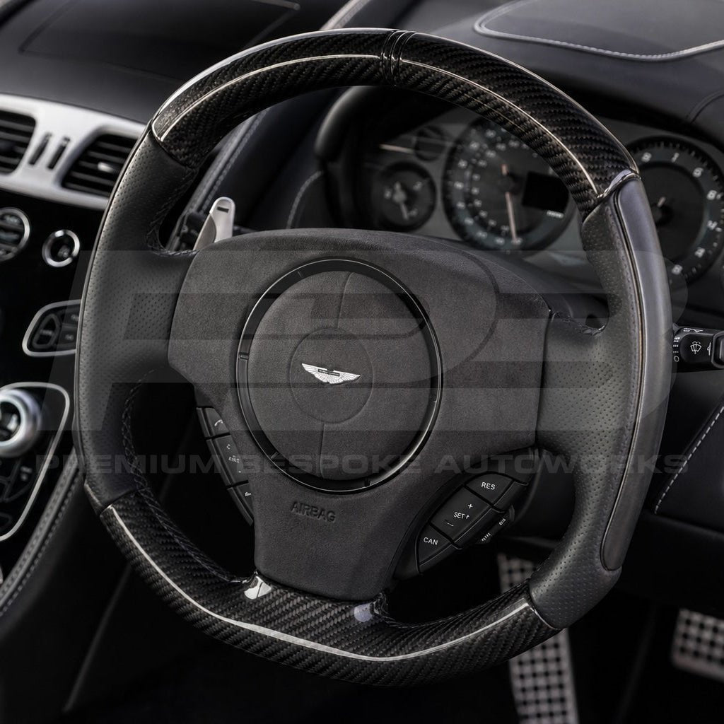 Close-up of a black carbon-fiber and leather Aston Martin steering wheel with the brand's wing logo in the center, set within a luxury car interior.
