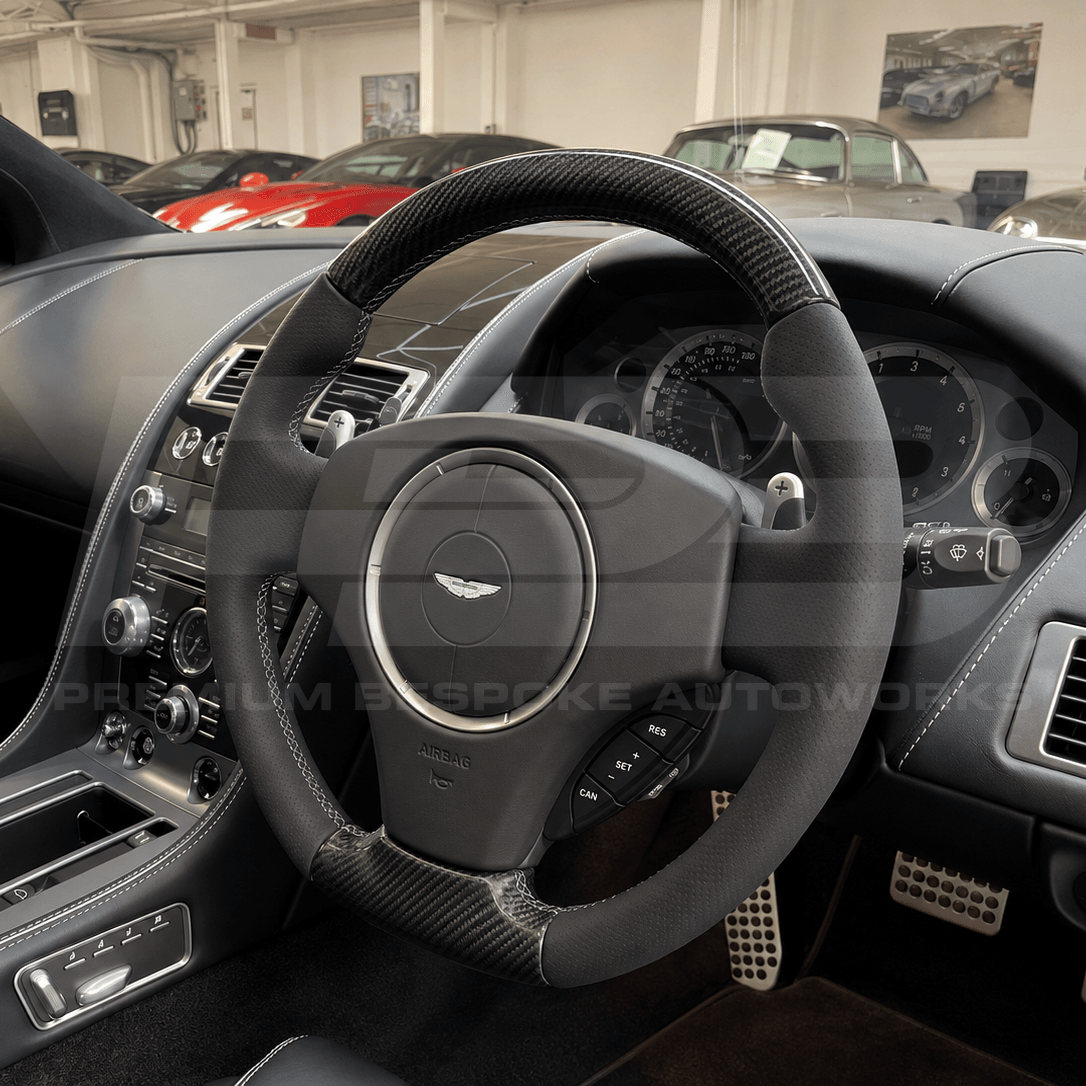 Close-up of a luxury car interior featuring a black leather and carbon-fiber steering wheel with the Aston Martin wing logo, a dark dashboard, and other vehicles visible in the background showroom.