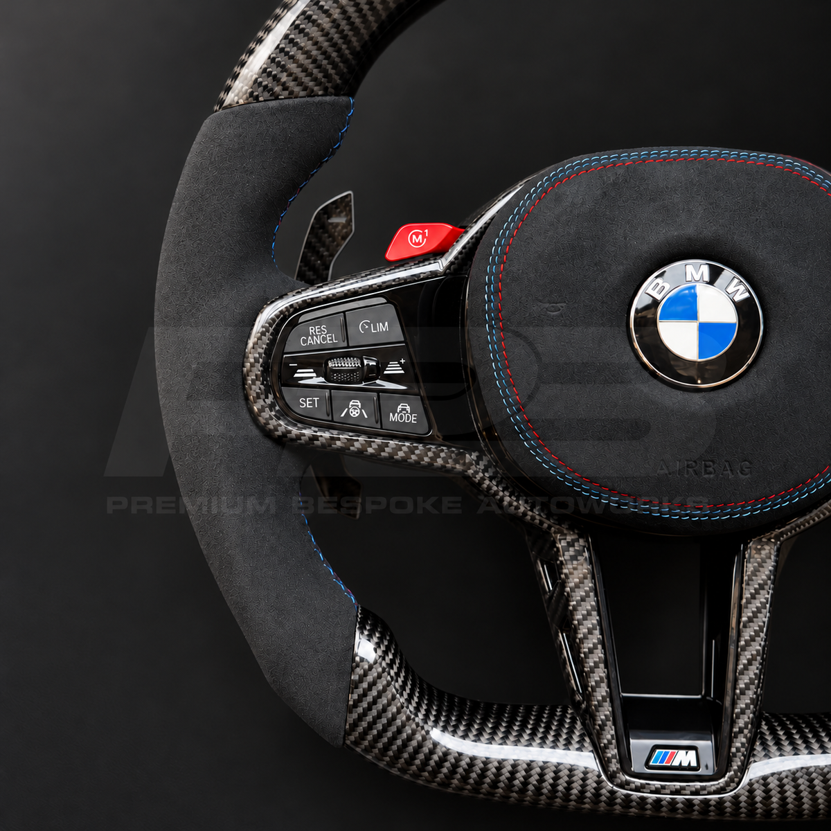 BMW M3 G80 LCI custom steering wheel close up detail showing flat bottom, gloss carbon fibre top, bottom and trims, black Alcantara grips and airbag cover with M tricolour stitching, red M buttons