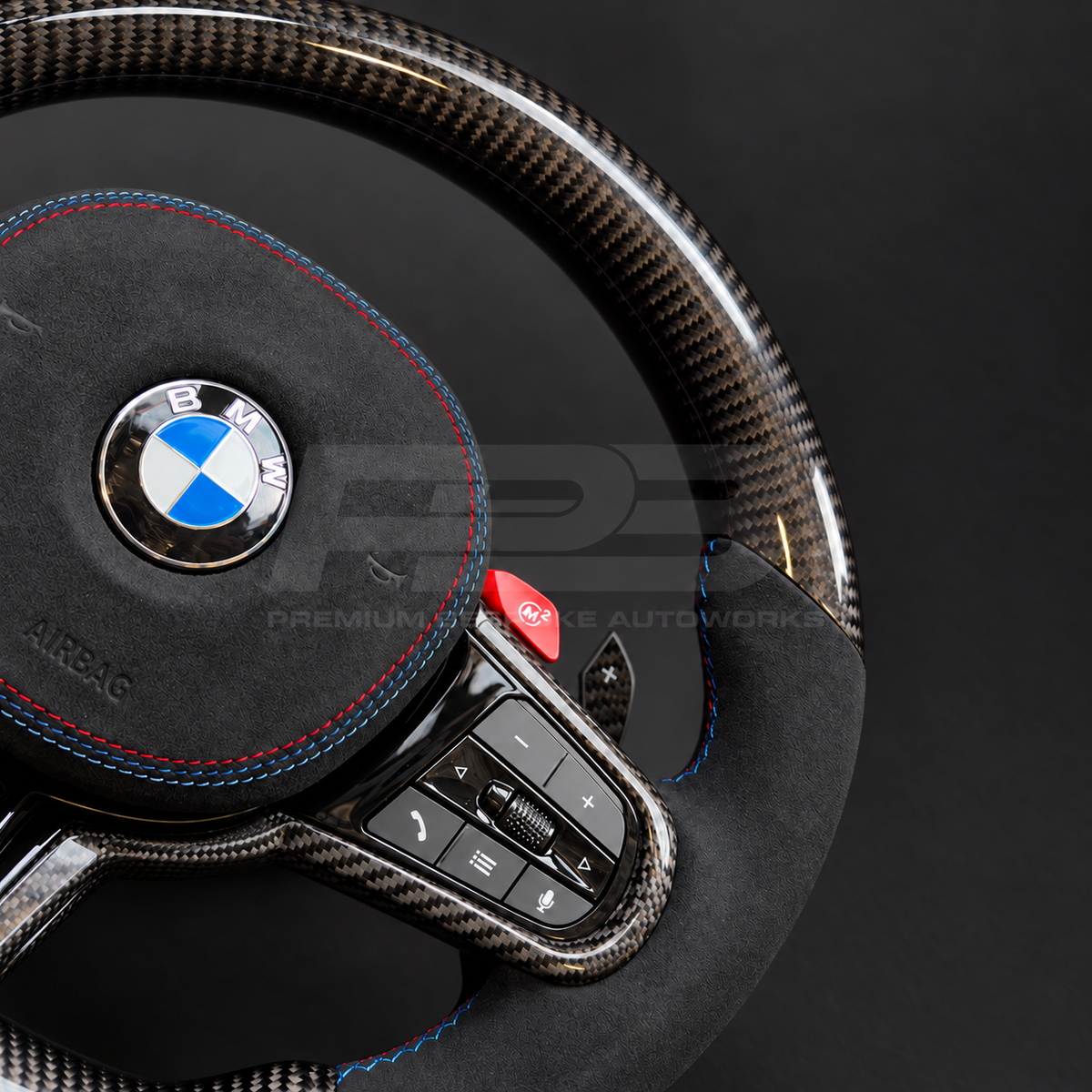 BMW M3 G80 LCI custom steering wheel close up detail showing flat bottom, gloss carbon fibre top, bottom and trims, black Alcantara grips and airbag cover with M tricolour stitching, red M buttons