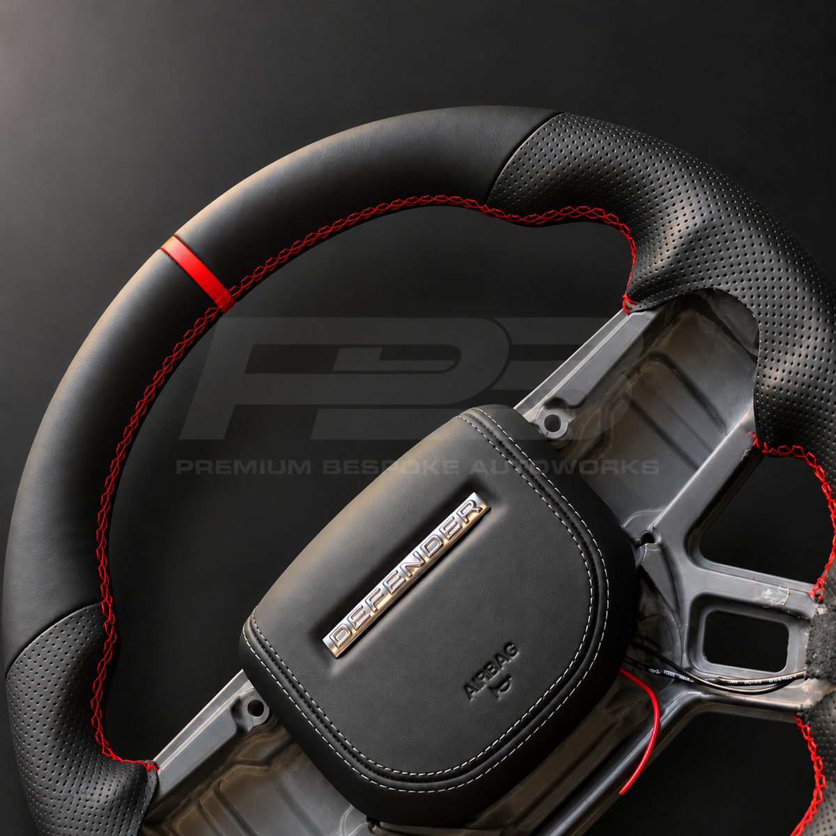 Land Rover Defender L663 custom steering wheel close up detail showing flat bottom, black nappa leather, perforated grips, red stitching and red marker, black leather airbag cover