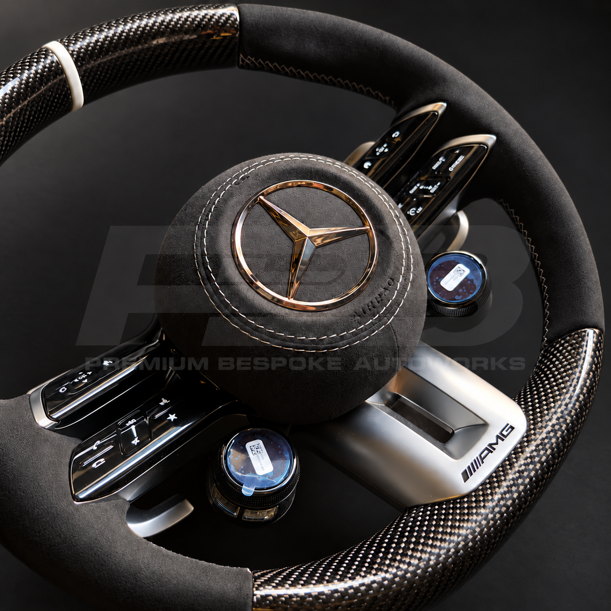 Mercedes-AMG G63 custom steering wheel close up detail showing gloss carbon fibre top and bottom, black Alcantara grips and airbag cover, classic grey stitching and marker, AMG drive unit dials