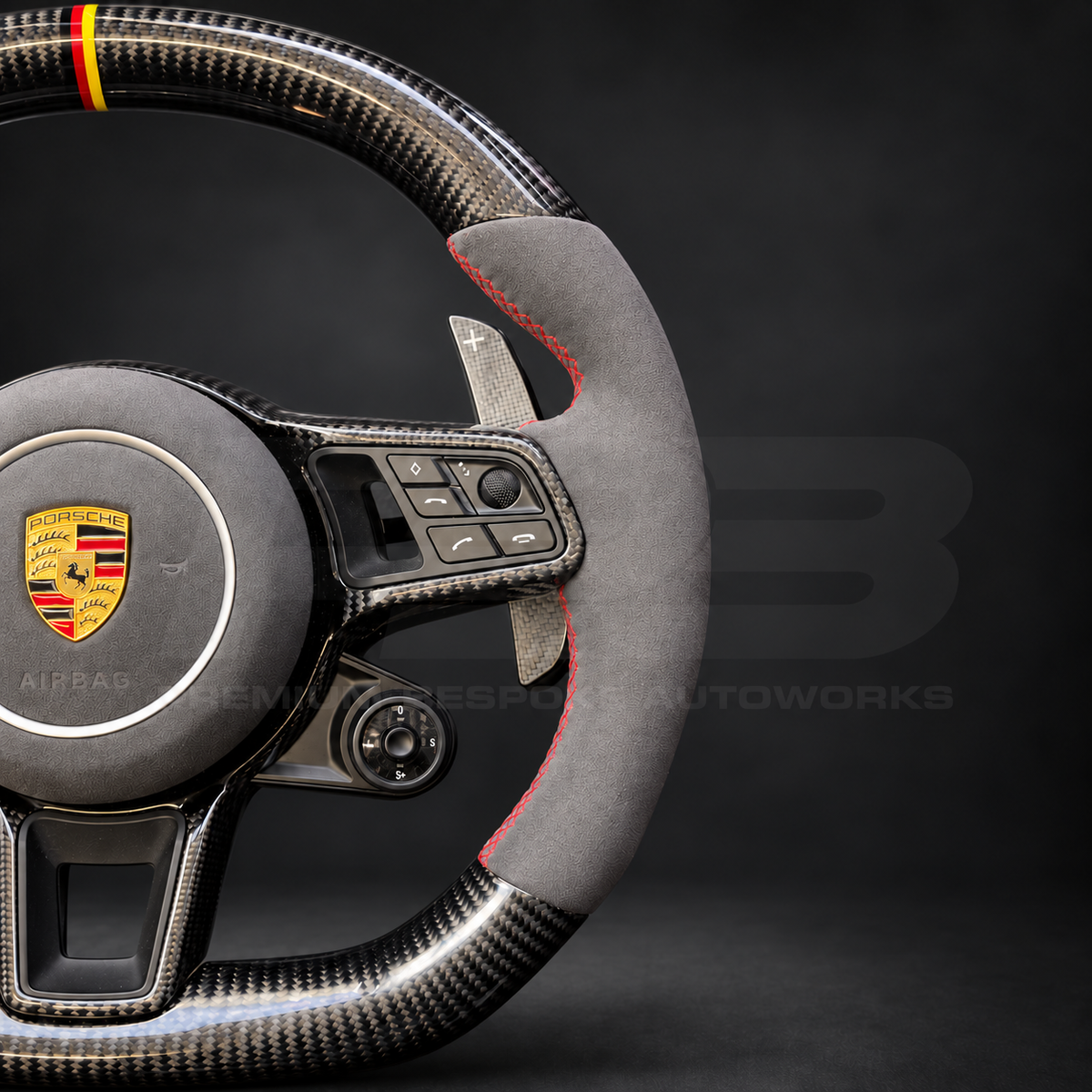 Porsche Cayenne 9Y0 custom steering wheel close up detail showing gloss carbon fibre, grey Alcantara grips and airbag cover, red stitching and German flag marker