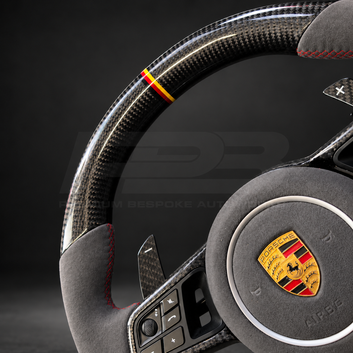 Porsche Cayenne 9Y0 custom steering wheel close up detail showing gloss carbon fibre, grey Alcantara grips and airbag cover, red stitching and German flag marker