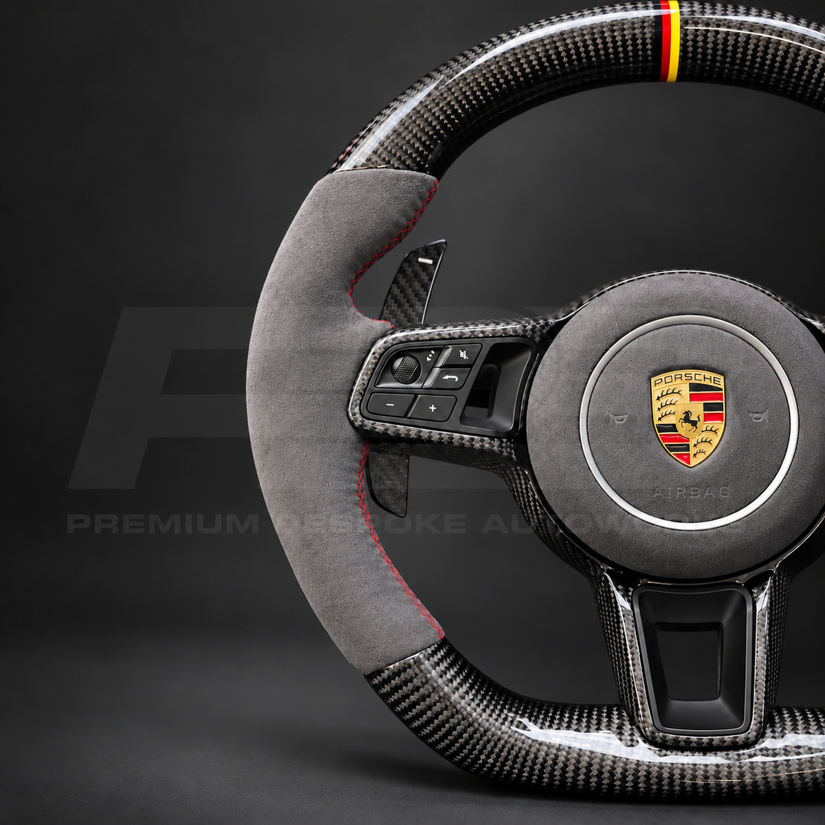 Porsche Cayenne 9Y0 custom steering wheel close up detail showing gloss carbon fibre, grey Alcantara grips and airbag cover, red stitching and German flag marker