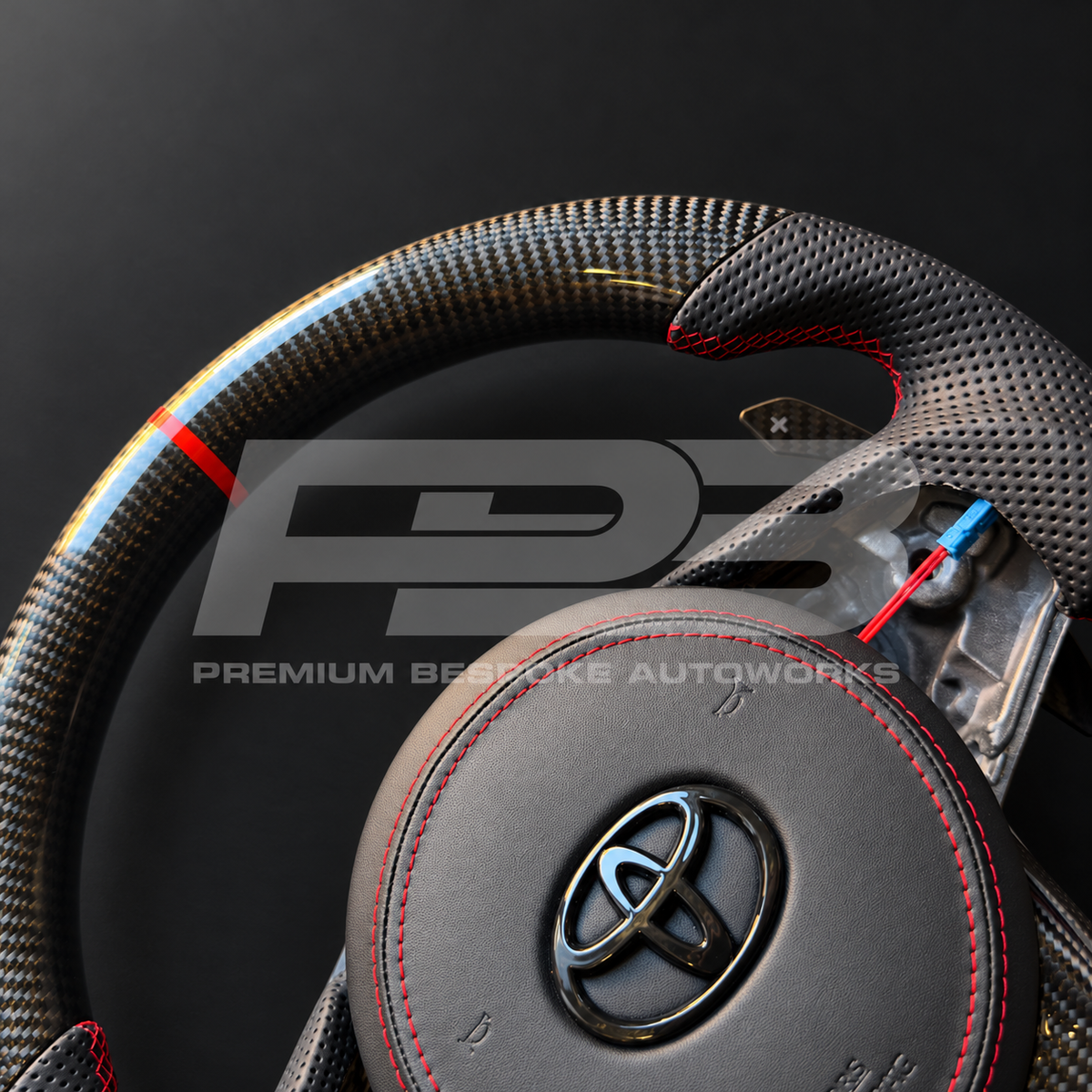 Close up of the gloss carbon fibre top section, red 12 o'clock marker, perforated leather grip and red stitched leather airbag cover on a custom Toyota GR Supra steering wheel
