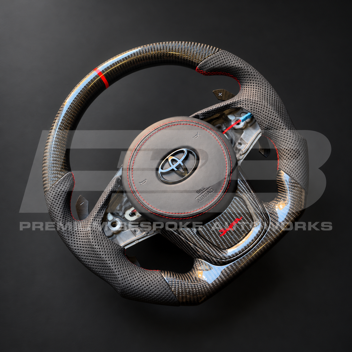 Toyota GR Supra MKV flat bottom carbon fibre custom steering wheel at an angle, showing the carbon lower spoke trim with red Supra script and magnetic paddle shifters